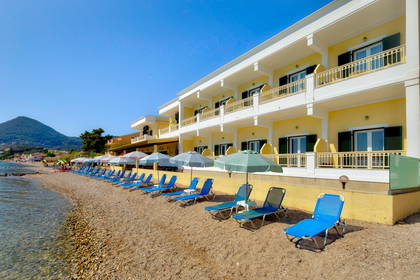 Żółty hotel z balkonami położony nad kamienistą plażą z leżakami i parasolami.