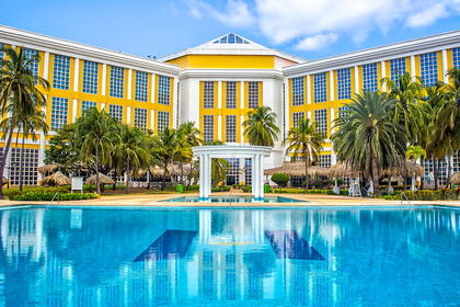 Hotel z basenem, palmami i błękitnym niebem w tle.