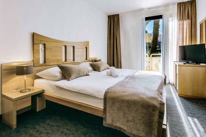 Jasny pokój hotelowy z podwójnym łóżkiem, szafką nocną z lampką, telewizorem i balkonem.