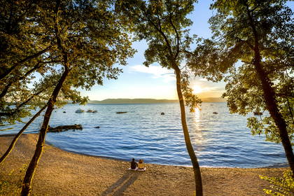 Dwie osoby na plaży patrzą spomiędzy drzew na zachód słońca nad jeziorem.