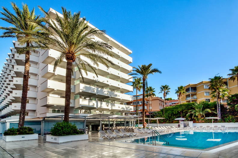 Biały budynek hotelowy z basenem otoczony palmami w słoneczny dzień.
