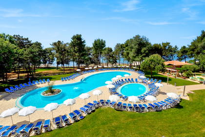 Kręty basen hotelowy z widokiem na morze, otoczony zielenią, przy którym stoją niebieskie leżaki i parasole.
