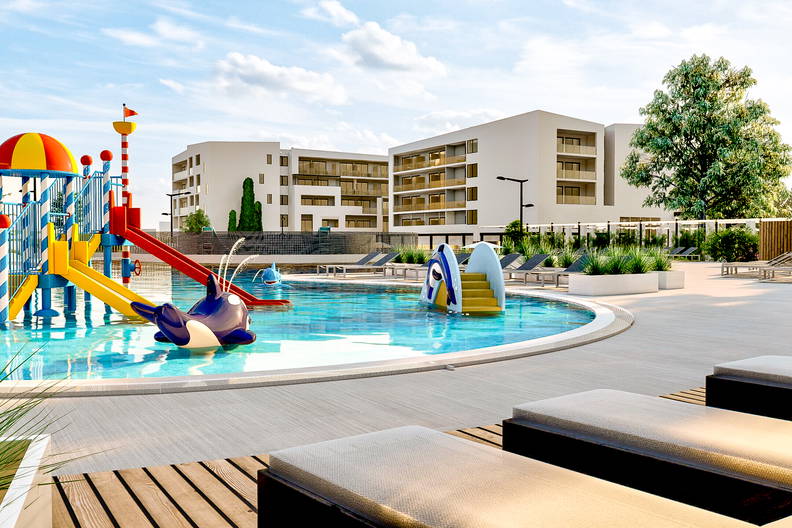 Wodny plac zabaw dla dzieci w ośrodku wypoczynkowym, z leżakami przy basenie i budynkami hotelowymi w tle.