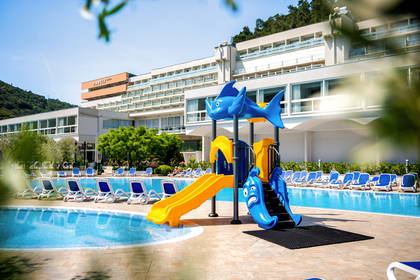 Kolorowy plac zabaw ze zjeżdżalnią przy hotelowym basenie otoczonym leżakami.