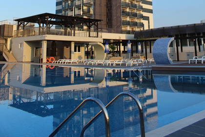 Wysoki hotel z basenem i leżakami, którego odbicie lustrzane widać w wodzie.