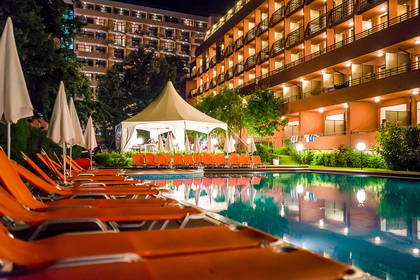 Basen hotelowy z leżakami oświetlony nocą. W tle pawilon.