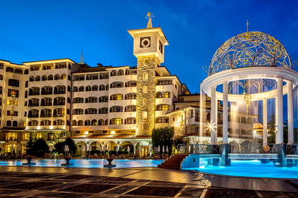 Hotel z wieżą zegarową i oświetloną altaną z fontanną nocą.