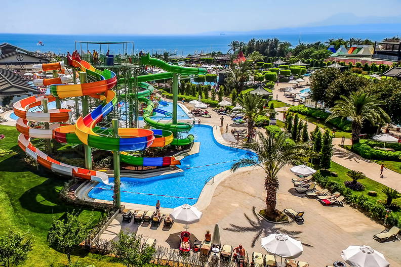 Aquapark z kolorowymi zjeżdżalniami, basenami, leżakami i palmami, widok z góry.