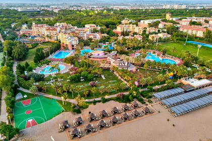 Widok z lotu ptaka na rozległy kompleks hotelowy przy plaży, z basenami, palmami i górami w tle.