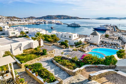 Widok z góry na białe budynki Mykonos, port, morze i góry w tle.
