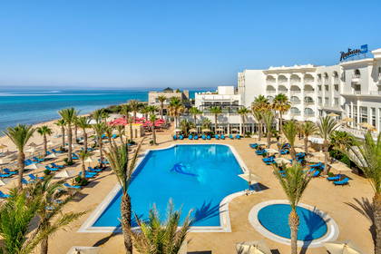 Hotel z basenem, palmami i plażą widziany z góry.