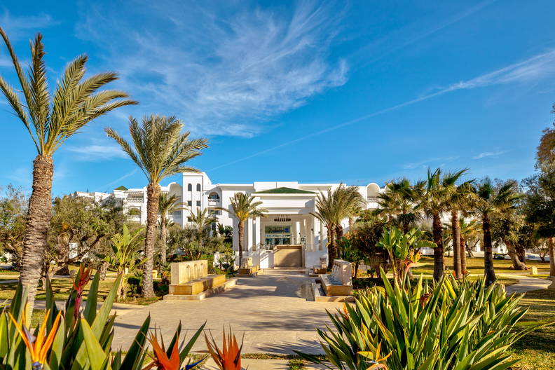 Biały budynek hotelu otoczony palmami i tropikalną roślinnością.