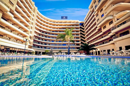 Basen hotelowy z leżakami i budynkami w tle.