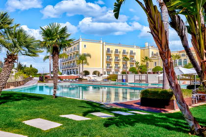 Żółty hotel z balkonami nad basenem otoczonym palmami.