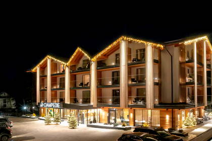 Oświetlony hotel nocą, widok z zewnątrz. Widoczne balkony i zaparkowane samochody.