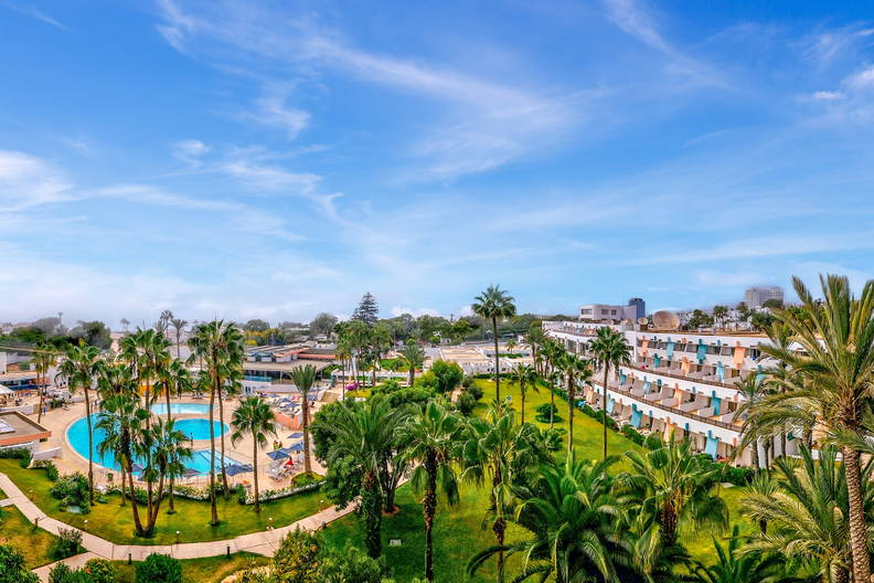 Panorama hotelu z basenem i palmami w słoneczny dzień.