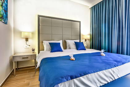 Pokój hotelowy z łóżkiem i niebieską pościelą. W oknach wiszą niebieskie zasłony.
