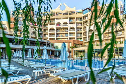 Kompleks hotelowy z basenem i leżakami widoczny przez gałęzie drzewa.