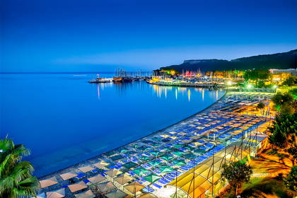Nocny widok na plażę, port z jachtami i oświetlone budynki.
