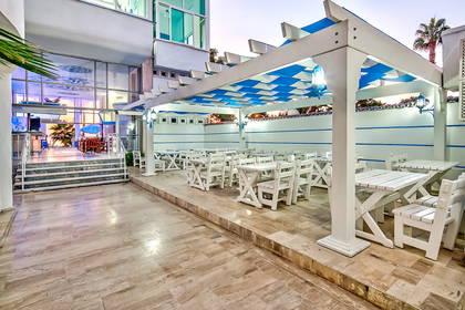 Białe stoliki i krzesła na tarasie restauracji z niebieską markizą.