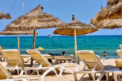 Słoneczna plaża z leżakami, słomianymi parasolami, turkusowym morzem i widoczną w oddali żaglówką.