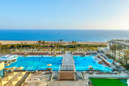 Kompleks hotelowy z dużymi basenami, leżakami i palmami, położony bezpośrednio przy piaszczystej plaży.