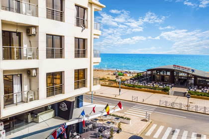 Hotel z kawiarnią i flagami w słoneczny dzień, widok na plażę i morze.