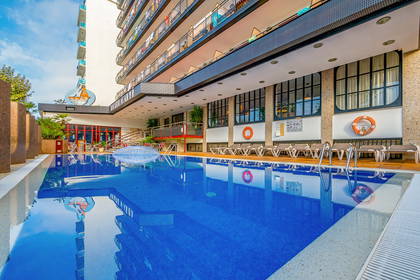 Basen hotelowy z leżakami, w którym odbija się budynek.