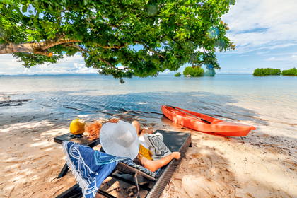 Kobieta w kapeluszu relaksuje się na leżaku na plaży. Obok stoi kajak.