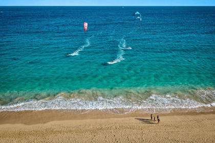 Plaża z kitesurferami na tle turkusowego oceanu i spacerujący ludzie.