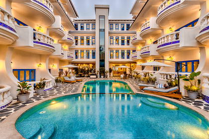 Hotel z białymi ścianami, niebieskimi oknami i balkonami przy basenie.