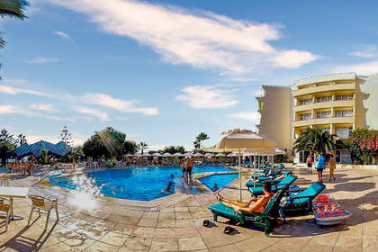 Panorama hotelu z basenem, palmami i leżakami w słoneczny dzień.