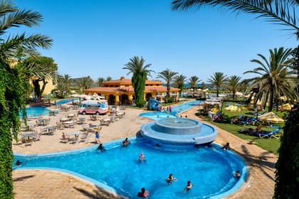 Kompleks hotelowy z basenami i palmami. W tle budynek hotelowy.