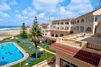 Hotel z basenem, leżakami na trawie i plażą w tle.