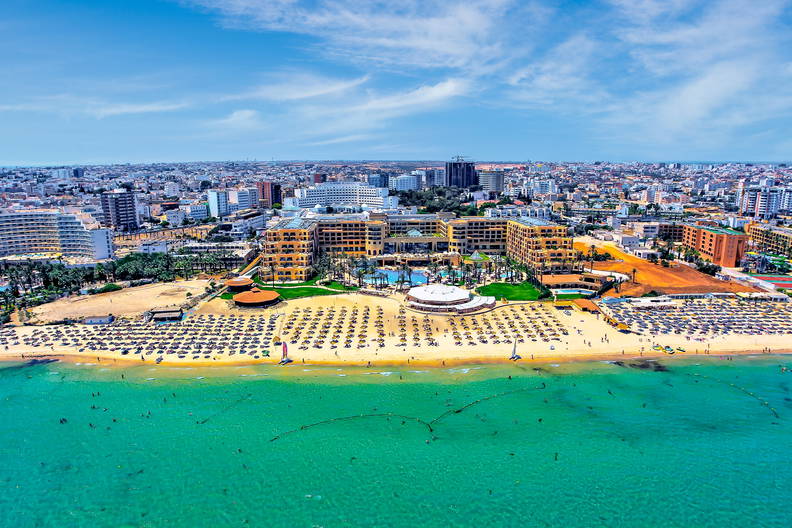 Plaża z turkusową wodą widziana z góry. W tle hotel i zabudowa miejska.
