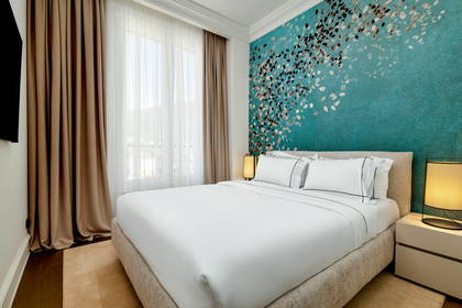 Sypialnia hotelowa z łóżkiem w białej pościeli na tle turkusowej ściany z motywem liści.