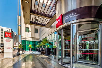 Wejście do hotelu Ibis Mall of the Emirates z obrotowymi drzwiami i roślinami.
