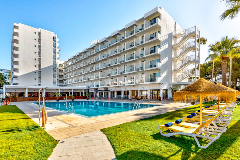 Duży, biały hotel z basenem i leżakami na trawniku.