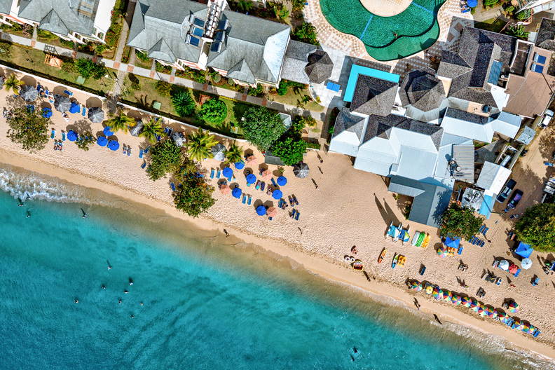 Plaża z turkusową wodą widziana z góry. Na piasku stoją parasole i leżaki, w tle widać budynki.