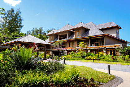 Hotel z szarą elewacją i dachem otoczony egzotyczną roślinnością.