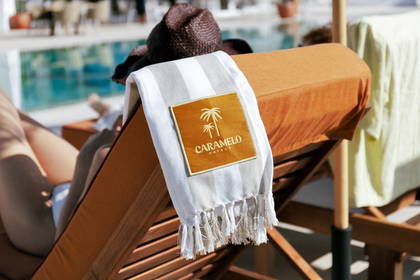 Osoba w kapeluszu wypoczywa na leżaku przy basenie. Na leżaku leży ręcznik z logo Caramelo Hotels.