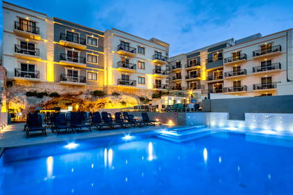 Oświetlony nocą hotel z basenem i leżakami.