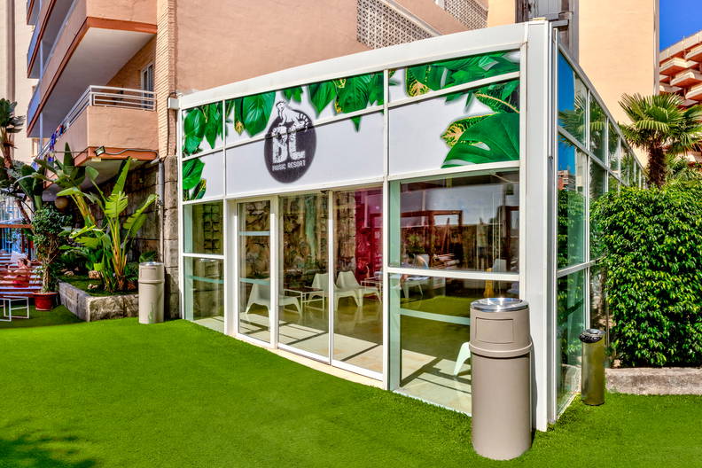 Pawilon z logo "BC Music Resort" i motywem roślinnym otoczony trawnikiem.