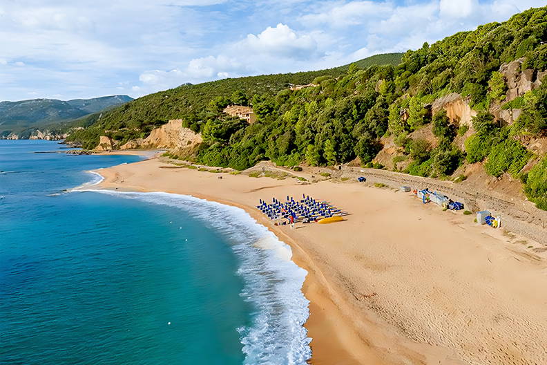 Plaża z błękitnymi parasolami widziana z lotu ptaka. W tle zielone góry.