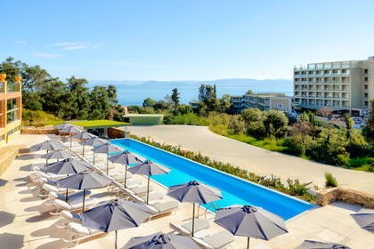 Basen hotelowy z leżakami i parasolami, widok z góry. W tle morze i budynki.