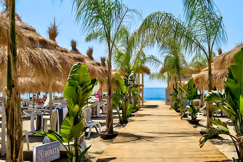 Drewniana ścieżka na plaży prowadzi do morza. Wokół palmy i parasole.