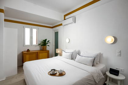 Pokój hotelowy w jasnych barwach z łóżkiem, szafką i rośliną.