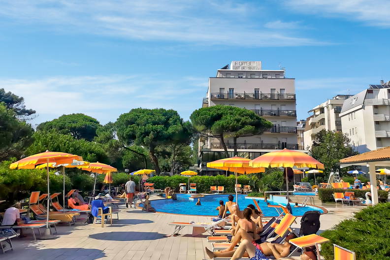 Basen hotelowy z leżakami i parasolami. Wokół wypoczywają ludzie.