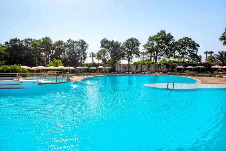 Basen w turkusowym kolorze otoczony zielenią i parasolami.