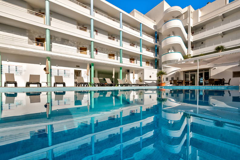 Basen z turkusową wodą przed hotelem w słoneczny dzień.